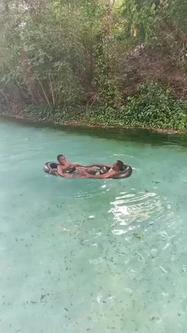 Two young people, likely children, are floating in a clear, turquoise-colored river or spring. They are each positioned inside a black inflatable tube, holding hands to stay together. The water is shallow enough to see the bottom, where there are rocks and some vegetation. The river is bordered by dense green trees and foliage. The atmosphere appears relaxed and peaceful.