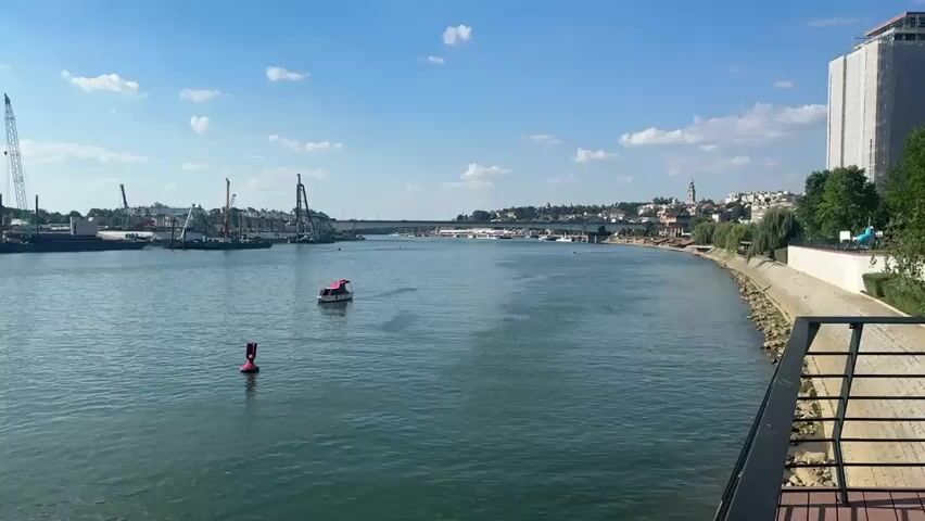 A wide shot captures a calm, blue river under a bright, partly cloudy sky. In the foreground, the right side of the frame shows a railing and wooden platform, hinting at a waterfront promenade or viewing area. The river stretches out towards the horizon, with a distant city skyline visible. On the left side of the river, a bustling industrial area is present, featuring several large cranes and barges, suggesting construction or port activity. A small red and white boat navigates the water in the middle ground, and a red buoy is visible closer to the camera. On the right bank of the river, a paved walkway lined with trees leads towards the city. A tall, modern building, partially covered in scaffolding, stands prominently on the far right. The overall atmosphere is one of urban development meeting natural waterways on a sunny day.
