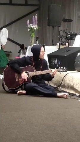 A person wearing a black hooded sweatshirt and glasses is sitting cross-legged on the floor and playing an acoustic guitar. They are also wearing a beaded necklace. In the background, there is a bouquet of flowers, a fan, a speaker, and what appears to be drum equipment. The lighting suggests an indoor event or performance space.