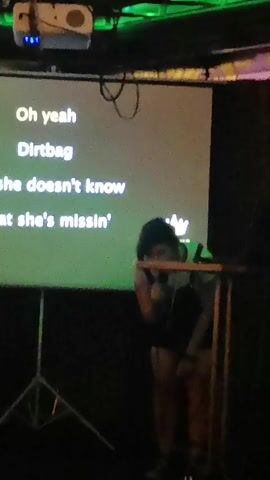 The video begins with a projection screen displaying lyrics that read "Oh yeah," "Dirtbag," and "she doesn't know," followed by "what she's missin'." A person wearing a dark shirt and shorts is singing into a microphone, leaning forward. They are standing on what appears to be a stage, with a projector visible in the upper left corner of the screen. The background is dark, with some green lighting. The scene then shifts abruptly. The subsequent frames are dark and blurry, making it difficult to discern specific details. There are glimpses of people and objects, but they are indistinct. One frame shows a red light. Another frame shows what might be a reflection in glass or a screen, with blurry figures in the background. The final frames are extremely dark and blurry.