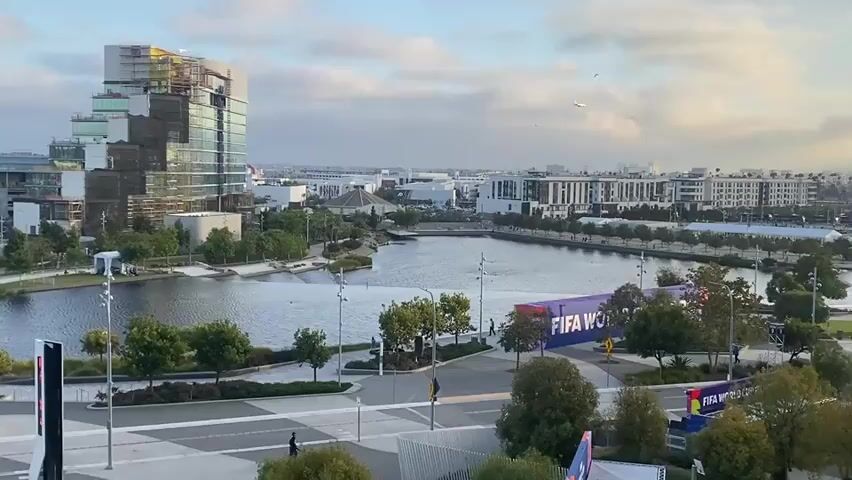 This is a panning drone shot of an urban area with a large body of water in the foreground. On the left side of the frame, there is a large, unfinished building under construction, with scaffolding and cranes visible. The building has a unique, stepped architectural design with glass and concrete elements. Across the water, on the right side of the frame, there are several buildings and structures, including what appears to be an entrance to a venue with large banners that read "FIFA WORLD CUP 2026". There are also trees and lampposts lining the waterfront. The sky is partly cloudy, with a few birds flying in the distance. A small airplane is visible in the sky towards the right side of the frame. In the lower foreground, there are paved walkways and some pedestrians are visible walking. The overall impression is of a modern, developing urban environment with an event possibly approaching, given the FIFA World Cup branding.