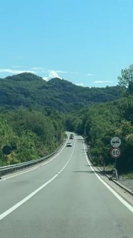 The video depicts a scenic daytime drive along a winding asphalt road through a mountainous and heavily forested region near Donji Ulići, Montenegro. The sky is bright blue with a few distant wispy clouds, indicating clear and sunny weather.

The road features a white dashed center line and solid white lines on the shoulders. Several cars are visible ahead, traveling in the same direction as the camera vehicle, which is moving forward. Dense green trees and lush vegetation cover the steep hillsides that flank the road, with some rocky outcrops visible among the foliage. Metal guardrails run along the left side of the road.

On the right side of the road, there are several traffic signs. A prominent circular sign with a red border displays "80," indicating a speed limit of 80 km/h. Above it, a triangular warning sign with a red border and a black symbol of two cars with a diagonal line across them signifies "no overtaking." Further ahead on the right, another triangular warning sign with a red border contains a black exclamation mark, indicating general danger or caution.

The bright sunlight suggests it is midday or early afternoon. In the background, music is audible, indicating it's playing within the vehicle from which the video is being recorded.