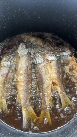 A close-up shot shows several small fish being fried in hot oil in a black pot. The fish are golden brown and appear crispy, with their fins and tails visible. Bubbles rise from the oil, indicating that the fish are actively cooking. The camera is positioned directly above the pot, offering a clear view of the food.