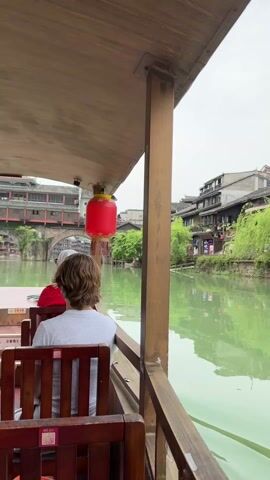 Here's a detailed description of the video:

The video shows a view from a boat traveling on a river in a town with traditional Chinese architecture. Two people are sitting in front, facing away from the camera. One person wears a grey shirt and the other a red one. The boat is covered by a simple wooden roof supported by wooden beams. A red lantern hangs from the roof. 

The river has a greenish hue. On both sides of the river, traditional buildings are visible, constructed with dark roofs and light-colored walls. A stone bridge with arches is seen ahead. Trees and green vegetation line the riverbanks. The sky is overcast.