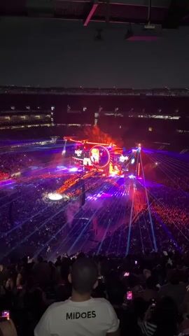 The video shows an aerial view of a large outdoor concert taking place at night. A massive crowd of people fills the stadium, illuminated by a spectacular light show featuring numerous lasers and spotlights in shades of purple and red, converging on a central stage. The stage is adorned with large screens displaying images of performers and is bathed in bright orange and red lighting. Smoke or fog drifts across the crowd and the stage. In the foreground, the back of a person's head wearing a white t-shirt with the word "MIDOES" is visible, obscuring the lower part of the frame. The surrounding stadium seating is also visible, with people watching the performance, many holding up their phones. The sky above is dark, indicating it is nighttime.