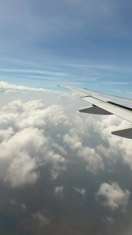 The video shows an aerial view from an airplane window. A portion of the airplane's wing is visible in the foreground on the right side of the frame. Below the wing and filling most of the frame are a vast expanse of white, fluffy clouds. The sky above is a clear blue with some faint wispy clouds. The ground is barely visible through gaps in the clouds, appearing as a dark, indistinct landscape with hints of man-made structures. The camera perspective suggests the airplane is in flight.