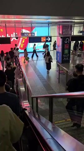 The video shows a public space, likely a subway station, with several people visible. On the left, an escalator leads down, with people ascending. In the middle ground, a group of people are walking across a tiled floor. Large digital billboards with Chinese text and graphics are prominent in the background. One billboard displays a red can with text that appears to be an advertisement. Another billboard shows a blue background with white text and a directional arrow. On the right, a sign indicates an exit and a route with arrows. The lighting is bright and artificial. People are dressed in casual clothing. Some individuals are carrying bags or backpacks. The overall atmosphere appears to be busy and functional, typical of a transit hub.