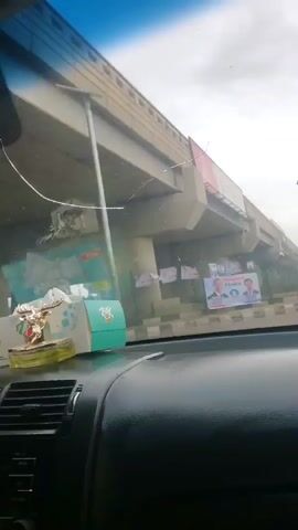 The video shows a view from inside a car, driving on a road in what appears to be a town or city. There is an overpass, with political posters attached to it. The car is driving behind another car. Pedestrians are crossing the street, and there is a yellow tricycle in the distance. The sky is overcast. There are street lights with solar panels, and buildings lining the road. The dashboard of the car is visible in the foreground. There is music playing in the background.