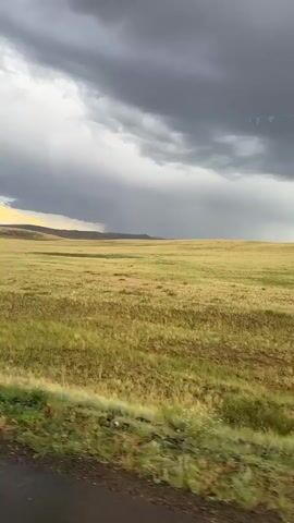 The video captures a vast, open landscape of rolling hills and a wide expanse of dry, golden grass under a dramatic, stormy sky. The foreground features a dark, wet road and a strip of vibrant green grass, suggesting recent rain or an ongoing shower. In the distance, the terrain slopes gently upwards, with faint outlines of mountains visible beyond the fields.

The most striking element is the sky, filled with heavy, dark grey storm clouds. A distinct shaft of rain is visible in the middle distance, falling from the clouds onto the grassy plain. The light filtering through a break in the clouds on the left creates a pale yellow glow, contrasting with the ominous dark clouds overhead. The overall mood is one of impending or ongoing weather, with a sense of wild, untamed nature.

The scene suggests a rural or remote location, possibly a steppe or prairie. The presence of the road indicates it might be a landscape viewed from a vehicle, possibly a car or bus. There are no visible people or man-made objects other than the road, emphasizing the natural elements of the scene.

The time of day is not precisely discernible but the quality of light, with the contrast between the dark storm and the brighter areas, suggests it could be late afternoon or early evening. The weather is clearly inclement, with a storm or heavy shower taking place. The location is identified as Бүркітті, Kazakhstan, which aligns with the description of a vast, open, and potentially dry grassland environment. There is no visible text in the media.