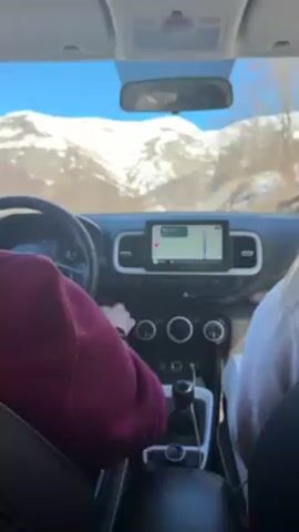 From inside a vehicle with two occupants, a view of snow-covered mountains is visible through the front windshield on a clear, sunny day. The driver, wearing a maroon sweater, is behind the steering wheel. The passenger, in a white sweatshirt, reaches towards the car's dashboard, near the infotainment screen which displays a navigation app. The car is equipped with a manual transmission, and a road sign is visible outside on the right side of the road.