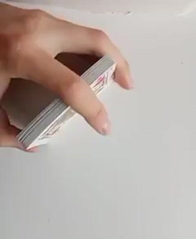 Two hands are shown shuffling a deck of cards. The cards are fanned out and rifled together. The shuffling is done with the backs of the cards facing the viewer, revealing a blue, ornate pattern on each card. The background is a plain white surface. The lighting is bright and even.