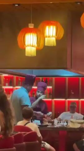 A video shows two chefs wearing purple hats cooking food on a large grill while a group of customers watch. The chefs work together, one preparing ingredients and the other using utensils on the grill. The customers sit around the grill, some watching intently while others talk and laugh. Red lighting illuminates the shelves behind the grill, displaying a variety of bottles and decorative objects. Overhead, three decorative lamps with orange shades hang from the ceiling. The scene appears to be a hibachi-style restaurant.