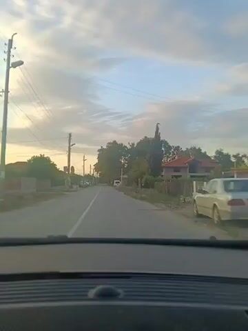 The video shows a point-of-view shot from inside a vehicle traveling down a paved road. The road is lined with trees and houses on both sides. There are utility poles with wires running along the left side of the road. The sky is cloudy with patches of blue. The time of day appears to be late afternoon or early evening due to the soft lighting. There are a few parked cars visible along the side of the road and a white van further down. The houses appear to be simple, residential structures. The overall impression is a rural or semi-rural setting.