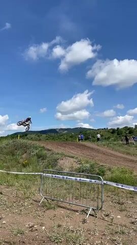 A motocross rider in a red and white suit on a white dirt bike navigates a dirt track under a bright blue sky with fluffy white clouds. The track curves around a grassy hill, and spectators are visible in the background. A metal barrier with white tape runs across the foreground, separating the track from the viewer's perspective. The overall scene depicts a motocross event on a sunny day.
