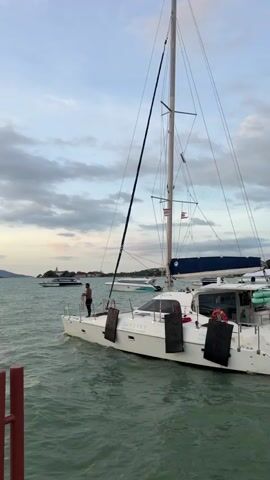 A white catamaran is docked in the water, with a person standing on the deck. There are other boats in the background, and the sky is cloudy. A small inflatable boat is seen moving away from the catamaran.