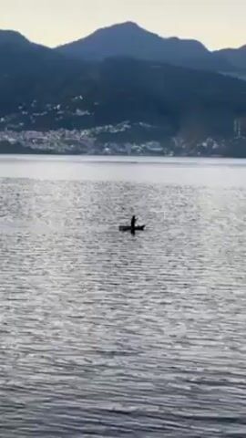 The video shows a single person standing on a small boat or paddleboard in the middle of a body of water. The water is rippled and gray. In the background, there is a shoreline with buildings and dark, mountainous terrain. The sky is overcast and pale. The person and the boat appear as silhouettes against the water. The overall tone is serene and quiet.