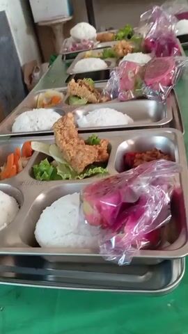 The video shows a close-up of a lunch tray, filled with various food items. The tray is divided into several compartments. In one compartment, there is white rice. Another compartment holds what appears to be fried chicken. There is also a serving of vegetables, including lettuce and carrots. A small portion of a reddish-brown dish is also visible. In the foreground, a dessert is packaged in clear plastic, revealing slices of dragon fruit. The tray is made of metal and has the inscription "RUSSO" on it. The tray is placed on a green surface, possibly a table. In the background, other similar trays filled with food are visible, arranged in a line. The lighting suggests an indoor setting, likely a cafeteria or dining hall. No people are clearly visible in the frame.