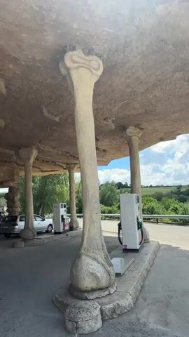 This video showcases an outdoor scene at a gas station with unique, bone-shaped pillars supporting a rocky overhang. In the foreground, a large, white, bone-shaped pillar with rounded ends supports the textured ceiling. Behind it, another similar pillar is visible. To the right, a white gas pump stands next to the pillar. In the background, a silver car is parked at another gas pump. Several people are visible near the car, appearing to be refueling or interacting. The sky is blue with scattered clouds, and trees and greenery surround the area, suggesting a rural or semi-rural location. The video pans up to reveal large, red, block-lettered signs attached to the rocky overhang, spelling out "GUST". The sun is shining brightly, casting a warm light on the scene.