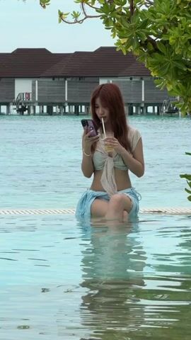 A woman sits by the edge of a swimming pool, holding her phone and a drink. She has auburn hair and is wearing a bra-like top, and a denim skirt. She is seated cross-legged, with her legs in the water. Her right leg crosses over her left. She has a straw in her mouth and is looking at her phone. Behind her is the ocean and some buildings on stilts. The water is clear and turquoise, and the sky is overcast. A tree branch hangs over the right side of the frame.