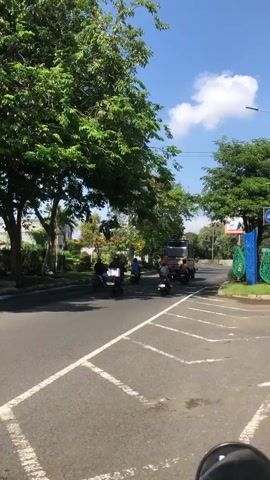 The video opens on a bright, sunny day, with a clear blue sky dotted with a few white clouds. The camera is positioned at street level, possibly from a vehicle, looking down a paved road. On the left side of the road, large, lush green trees with full canopies create a dense shade. Beyond the trees, a glimpse of a park-like area with more trees and some decorative structures can be seen. Several people are riding motorcycles on the road, moving away from the camera. One truck is also visible, further down the road, traveling in the same direction as the motorcycles. On the right side of the road, there are more trees and some colorful decorative elements, possibly public art or landscaping. The road itself is marked with white lines, indicating lanes and possibly a turn or intersection ahead. The overall scene depicts a typical urban or suburban street with moderate traffic and abundant greenery.