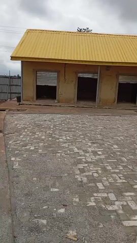 The video pans across an outdoor area with a building that has a yellow roof and several open doorways. The ground is paved with interlocking bricks and gravel. A hose lies on the ground. There is a tire visible in front of one of the doorways. The sky is overcast.