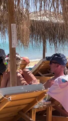 Two people are relaxing on lounge chairs under a thatched umbrella at a beach. The person on the left, facing away from the camera, wears a light-colored baseball cap and appears to be sunbathing. The person on the right, also facing away, wears a dark baseball cap and sunglasses, and is wearing a pink shirt. They are seated at a wooden table with drinks. In the background, the ocean is visible, and a person in a black swimsuit is walking on the beach. The scene is illuminated by bright sunlight.