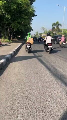 The video shows a street scene in Klurak, Indonesia, likely during the daytime due to the bright sunlight and long shadows cast on the asphalt road. The weather appears to be clear and sunny. The scene is a road with multiple motorcycles being ridden.

The primary subjects are people riding motorcycles. In the foreground, a person wearing a bright green helmet and a green jacket is riding a motorcycle with a large brown box secured to the back. Behind them, another person wearing a white shirt and a black helmet is riding a motorcycle. Further down the road, another individual on a motorcycle can be seen, and a black SUV is also present, along with more motorcycles in the distance. The road itself is paved but appears somewhat worn, with a concrete curb on the left side. To the left of the road, there is a large tree providing shade. In the background, there are some buildings and palm trees, suggesting a semi-urban or suburban environment.

The activity taking place is the transit of vehicles on a road. The presence of a large box on the motorcycle in the foreground suggests a delivery or transport of goods. The overall mood is one of daily life and movement. There is no visible text in the video.