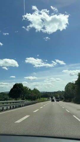 The video shows a first-person perspective of a car driving on a highway on a sunny day. The sky is blue with scattered white clouds. Trees line the side of the road. There are other cars on the highway. A speed limit sign of 50 is visible on the left side of the road. The car appears to be moving at a moderate speed.