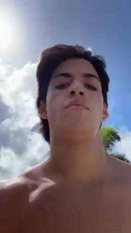 A shirtless young man films himself walking down a sunny street on a sidewalk.  There are houses, parked cars, trees, and grass visible.  The sky is blue with fluffy white clouds.  At one point, he holds up his hand, making a finger gun gesture to the sky.