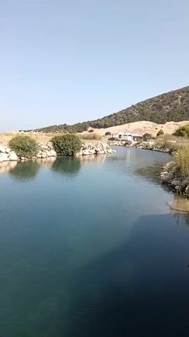 A wide shot captures a tranquil body of water with clear blue hues. The water's surface is mostly calm, reflecting the clear, bright sky above. On the left bank of the water, rocky outcrops are visible, interspersed with patches of dry grass and low-lying green bushes. Further back, a gently sloping hill rises, covered in sparse vegetation and dry earth. A few buildings, appearing to be small houses or structures, are nestled on the side of the hill.

The right bank of the water features more rocks and taller, dry grass. Towards the foreground on the right, a wooden structure, possibly a small dock or platform, extends over the water. A makeshift shelter with a green tarp is set up on this platform, supported by concrete blocks and wooden planks. Some objects are visible under the tarp, including what appears to be a white conical object, possibly a seating element or a decorative piece, and other miscellaneous items. The overall scene suggests a peaceful, rural waterfront setting on a sunny day.