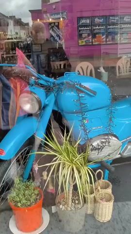 The video pans across a shop window displaying two vintage scooters. The scooter on the left is bright blue and decorated with small white string lights. It has a wicker basket on the rear with some red items inside. The scooter on the right is white and classic in design, with a brown seat and also a wicker basket on the rear, this one containing what appears to be a white pot or bowl. Several smaller wicker baskets are also visible in front of the scooters and inside the shop. Behind the scooters, shelves are visible stocked with various products and there are chairs and tables suggesting a seating area. A person is partially visible in the reflection on the window to the left, appearing to be a man with light-colored hair. The overall impression is a quaint and decorative shop display.