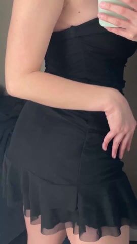 A person in a black, strapless mini-dress takes a selfie in front of a mirror. The dress has a ruffled hem. The person holds a light green smartphone in their right hand. The mirror is in what appears to be a dressing room or closet with a light-colored wall. A pile of clothes is visible in the background. The person shifts their body slightly while holding the phone.