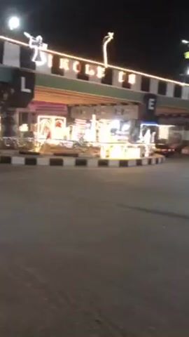 The video shows a street scene at night in what appears to be a city. A bridge or overpass spans the road, with illuminated lettering visible, partially spelling out "BIKOLE". The structure is decorated with lights and possibly figurines. A motorcycle is seen driving on the street. There are streetlights illuminating the area, and a traffic signal is displaying a red light. There are also billboards visible near the traffic signal. The overall scene seems to be depicting a public space decorated for a festive occasion.