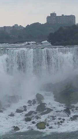 The video captures a wide shot of a powerful waterfall. Water cascades over a rocky ledge, creating a dense mist that partially obscures the lower portion of the falls and the rocks below. In the background, buildings and trees are visible on a higher elevation, suggesting an urban or semi-urban setting near the natural landmark. The sky appears overcast. The timestamp indicates the video begins at 00:00 and ends at 00:05. No people or specific interactions are discernible.