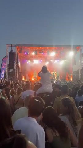 The video shows a concert with a live band performing on a stage. The stage is set up outdoors with lighting and speakers. A large crowd of people are gathered in front of the stage, many holding up their phones to record the performance. One person is being carried on someone's shoulders in the crowd, filming the band. The lighting on stage is a mix of red and purple. The sky appears to be dusk or evening.