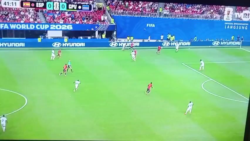 A soccer game is being played between two teams, one in red and the other in white. The score is tied at 0-0, and the game clock shows 41:14. The players are on a green grass field with white lines marking the boundaries. There are spectators in the stands in the background, wearing red shirts. A large screen displays the FIFA World Cup 2026 logo and the words "OFFICIAL BROADCASTER LANGSUNG". There are advertisements for Hyundai around the field. The players are in motion, with some running with the ball and others defending. One player in white is on the ground, possibly injured.