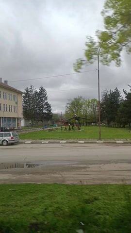 The video shows a static display of a military jet in a public park in a rural area. The jet is painted green and mounted on a pole, giving the impression it is in flight. The park is grassy with a sidewalk and a street. There are some trees around the edge of the park. The sky is overcast. The video appears to be taken from a moving vehicle.
