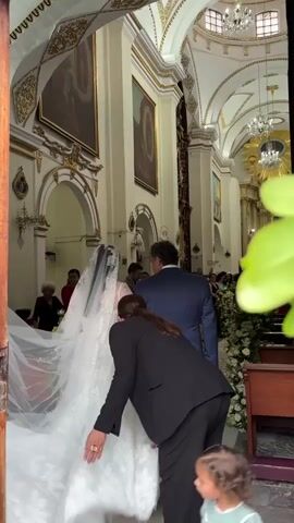 The image captures a significant moment inside a grand, ornate church, likely in Orizaba, Mexico. The scene depicts a wedding ceremony, with the bride about to enter or proceeding down the aisle.

In the foreground, the bride is seen from behind, wearing a voluminous white lace wedding gown with a long, flowing train and an intricate lace-edged veil covering her dark hair. To her right, a man in a dark blue suit stands beside her, also viewed from the back, likely the groom or her father, accompanying her. A woman in a black pantsuit, with her dark hair in a ponytail, is bending down to meticulously adjust the bride's extensive train, indicating her role as an attendant or wedding planner ensuring the dress is perfectly arranged. A small child is partially visible in the bottom right corner, looking upwards.

The church interior is distinguished by its high ceilings, white walls adorned with ornate gold trim on arches and columns, and large framed religious paintings hanging prominently. Chandeliers are visible overhead, and the aisle is decorated with white floral arrangements and green foliage. Other guests are blurred in the background, some seated in pews and others standing near the entrance. The atmosphere is one of anticipation and ceremonial preparation, as the bride is poised for her walk down the aisle.