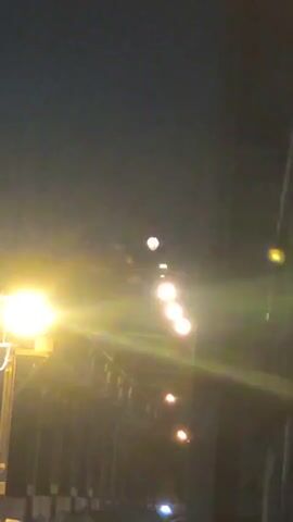 The video shows a nighttime view of a street or walkway lined with lights. The lights are yellow and appear to be street lamps or similar fixtures. Some of the lights are brighter than others, and there is a strong glare from the light on the left side of the frame. The background is dark, suggesting it is nighttime or indoors with minimal ambient light. There are some indistinct structures visible below the lights. The camera is angled upwards towards the lights and the sky.