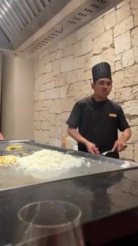 This is a short video showing a chef in a black uniform and chef's hat, with a yellow logo on his chest, standing behind a teppanyaki grill. He is holding cooking utensils and appears to be talking or performing for guests, as indicated by his animated gestures and facial expression. In the foreground, a person in a white shirt is visible, looking down and out of frame. The background features a stone wall. The video appears to capture a moment of a teppanyaki performance.