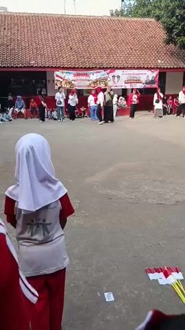 The video captures an outdoor gathering in what appears to be a schoolyard. In the foreground, two children are partially visible, facing away from the camera. One child wears a white hijab and a red shirt with a graphic design on the back. The other child's face is not visible, but they are wearing a red and white striped shirt.

In the mid-ground, several adults and children are engaged in activities. A group of adults, mostly women wearing red shirts, are standing and interacting. One woman in a green hijab is in the center, seemingly leading an activity or demonstration. Other adults are positioned around the yard, some observing, others participating. There's a banner in the background with Indonesian text and imagery, suggesting a national celebration or event. The ground is paved with a rough, light-colored surface, and a red-brick building with a tiled roof forms the backdrop. Trees are visible behind the building. Some children are seated on the ground or on the steps of the building. The atmosphere seems festive and communal.