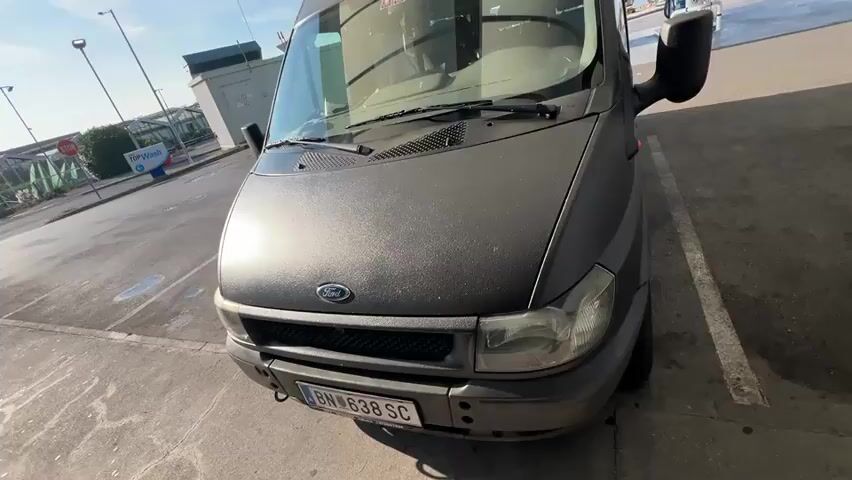 A dark gray Ford Transit van is parked at a gas station under a canopy. The van's license plate reads BN 638 SC. The van is angled towards the camera, with its front end visible. A gas pump is partially visible to the right of the van. The ground is asphalt, and there are white parking lines. In the background, there is a building with a blue and green sign, and the sky is blue with some clouds and the sun. The video appears to be taken from a handheld camera, possibly a smartphone. The perspective shifts slightly throughout the clip, suggesting the person filming is walking around the van.