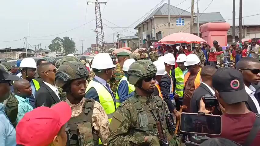 The video shows a crowd of people in an urban area.  Many are wearing white hard hats and some are in camouflage uniforms with helmets.  Several of those in uniform appear to be armed.  There are also people in suits and some in high-visibility vests. In the background, there are buildings, a utility pole, and a few parked vehicles.  The crowd appears to be moving slowly, possibly following someone. The weather looks overcast.