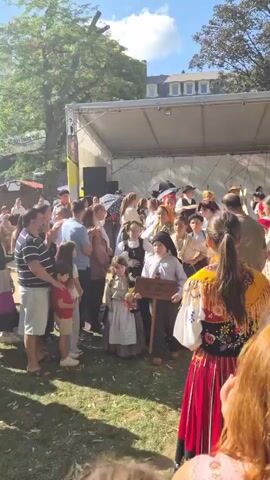 The video shows a group of people in traditional clothing on a sunny day outdoors. There is a stage set up in the background with a white canopy. In the foreground, a group of children dressed in historical attire, some holding bouquets of flowers, are walking towards the stage. One child in the center holds a sign on a stick that reads "Alta Nao". Adults are standing around watching and some are clapping. The environment appears to be a festival or cultural event.