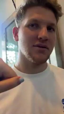A close-up video shows a man wearing a white shirt. Someone places their hand in front of his mouth. The hand has light blue nail polish and a gold watch. The man’s mouth is briefly covered by the hand. He then smiles. There is a logo for Wild Cherry on the man's shirt. He seems to be indoors.