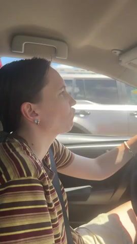 The video shows the interior of a car on a sunny day. A person with short, dark hair is driving, wearing a striped shirt and shorts. They are looking forward and seem to be talking or making a facial expression. The dashboard is visible, decorated with several small figurines, including a stuffed bear, a blue character, a pink hat, and a yellow character. A phone mount is also attached to the windshield. Outside the car, other vehicles are visible, and the sky is blue with some clouds. The overall scene suggests a casual drive.