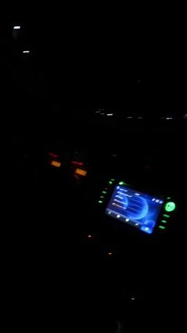 The video shows the view from inside a car driving on a snowy road at night. The car's dashboard is visible, with a lit-up speedometer and a digital screen displaying navigation information. The car's headlights illuminate the road ahead, revealing the snow-covered surface and surrounding vegetation. Distant streetlights and building lights can be seen. Music is playing inside the car.