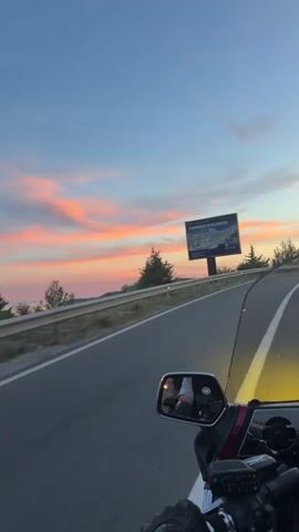 The video begins with a view of a billboard on the right side of a road, with trees and a hilly landscape in the background. The sky is a gradient of blue and pink, indicating either sunrise or sunset. A motorcycle is in motion, with the camera positioned behind the handlebars and looking forward. A side mirror reflects the rider and the sky. The road is paved with white lines. As the video progresses, the motorcycle continues to move along the road. A silver car passes by on the left lane. The camera continues to capture the road ahead and the scenic sky. The video ends with a similar view of the road, the sky, and the surrounding landscape.