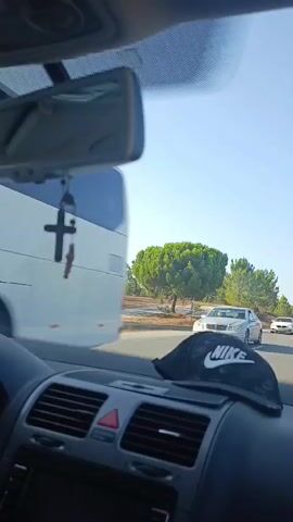 The video is shot from inside a car looking forward. The dashboard is visible in the foreground with a black Nike hat resting on it. Through the windshield, a road stretches ahead lined with trees. Several cars are visible on the road, including a white car in the middle distance and other vehicles further ahead. The sky is clear and blue. The car appears to be in motion.
