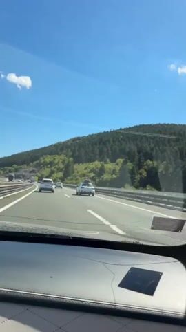 The video shows a view from inside a car driving on a highway. The highway has multiple lanes and is bordered by a guardrail. On the right side of the highway, there is a lush green hillside covered in trees. The sky is a clear blue with a few white clouds. Several cars are visible on the highway, including a white SUV with a roof rack, a white hatchback, and a darker colored vehicle further ahead. The car from which the video is recorded has a dashboard visible in the foreground, with a diamond-stitched pattern. The sun is shining, indicating it is daytime. The overall impression is of a scenic drive on a sunny day.